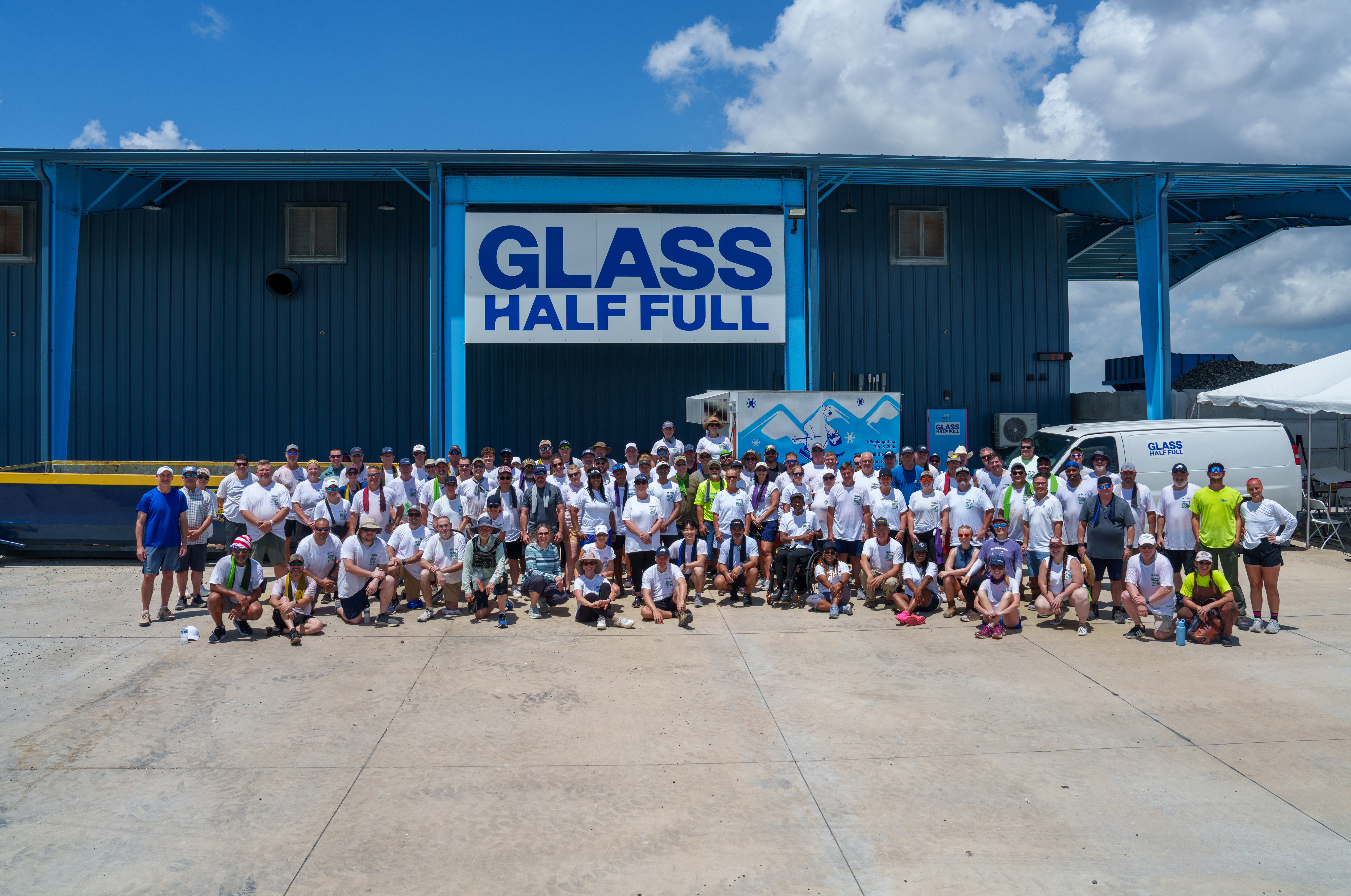 A group of around a hundered people in front of a blue steel building with a sign that reads Glass Half Full