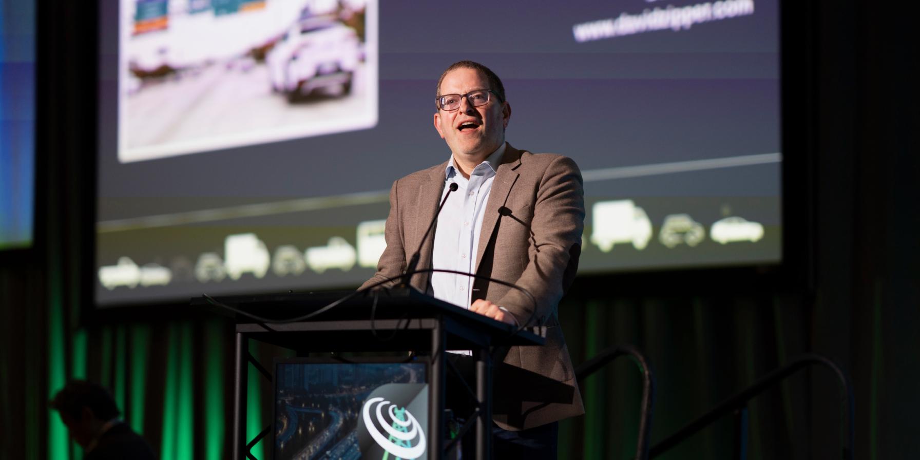 David Zipper speaking from a podium with presentation on large screen behind him