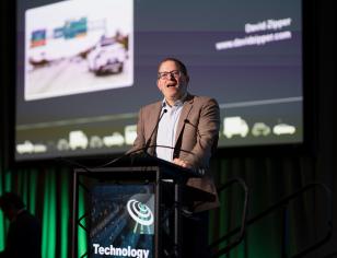 David Zipper speaking from a podium with presentation on large screen behind him