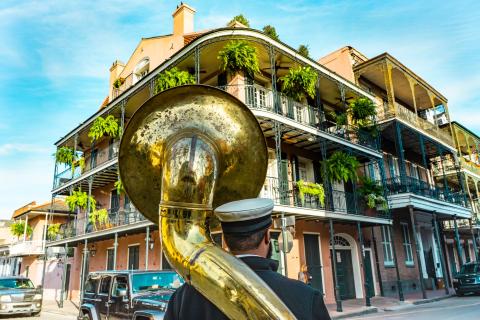 Historic French Quarter building in New Orleans with ornate wrought-iron balconies and hanging ferns, viewed behind a street musician carrying a large brass sousaphone.